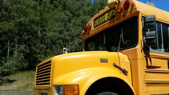 A classic yellow school bus is parked in a wooded area. The bus is large, with distinct black lettering on a sign above the windshield, and it has several lights on its top. The surrounding background consists of lush, green trees on a sunny day.