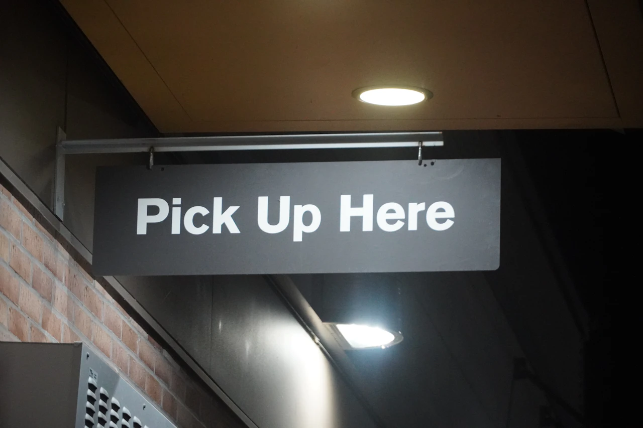 A Pick Up Here sign above a collection counter, where a reserved online order is handed over after an ID check