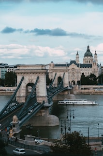 A historic stone bridge stretches across a river, with intricate metal railings and architectural details. A riverboat glides underneath, and a large building with a dome and spires is visible in the background. The scene is set under a partly cloudy sky.