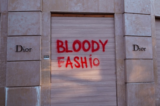 A storefront with boarded-up windows and the brand name 'Dior' visible on either side. The words 'BLOODY FASHION' are spray-painted in red across the wooden paneling covering the central display area.