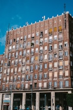 A multi-story brick building with numerous windows and balconies, some with metal railing and plants. The building features a mix of open, closed, and shuttered windows. A clear blue sky is visible overhead, and there is a store named Costa at the bottom of the building.