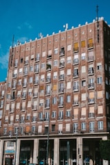 A multi-story brick building with numerous windows and balconies, some with metal railing and plants. The building features a mix of open, closed, and shuttered windows. A clear blue sky is visible overhead, and there is a store named Costa at the bottom of the building.