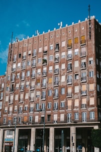 A multi-story brick building with numerous windows and balconies, some with metal railing and plants. The building features a mix of open, closed, and shuttered windows. A clear blue sky is visible overhead, and there is a store named Costa at the bottom of the building.