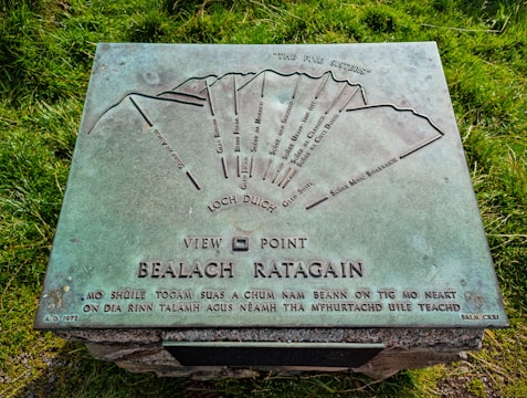 A metal plaque indicating various mountain peaks, featuring a detailed diagram with their names, orientation marks, and labels such as 'The Five Sisters.' The text at the bottom includes a Gaelic inscription, the location 'Bealach Ratagain,' and the year 'A.D. 1972.' The plaque is mounted on a stone structure surrounded by green grass.
