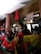A group of people are gathered, holding flags and signs. One person is holding a sign that reads 'STOP À LA MAFIA COLONIALE.' The scene takes place in a building with pillars and some people are capturing the moment with their phones.