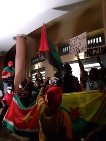 A group of people are gathered, holding flags and signs. One person is holding a sign that reads 'STOP &Agrave; LA MAFIA COLONIALE.' The scene takes place in a building with pillars and some people are capturing the moment with their phones.