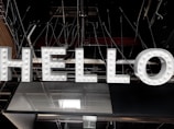 Large illuminated letters spelling out the word 'HELLO' are suspended against an industrial ceiling with visible beams and wiring. The letters are white with small light bulbs embedded in them, giving a classic marquee sign appearance.