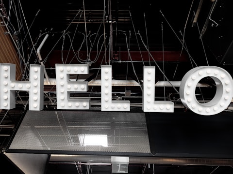 Large illuminated letters spelling out the word 'HELLO' are suspended against an industrial ceiling with visible beams and wiring. The letters are white with small light bulbs embedded in them, giving a classic marquee sign appearance.