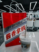 A red and white paper bag with stylized text in Chinese characters, featuring a graphic design with wavy patterns. A plastic cup with a straw, containing a layered iced beverage, is placed on a white table beside it. The background shows an interior setting with modern geometric ceiling lights and white chairs.