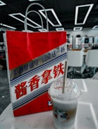 A red and white paper bag with stylized text in Chinese characters, featuring a graphic design with wavy patterns. A plastic cup with a straw, containing a layered iced beverage, is placed on a white table beside it. The background shows an interior setting with modern geometric ceiling lights and white chairs.