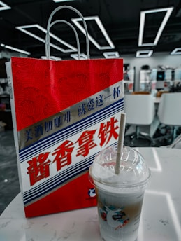 A red and white paper bag with stylized text in Chinese characters, featuring a graphic design with wavy patterns. A plastic cup with a straw, containing a layered iced beverage, is placed on a white table beside it. The background shows an interior setting with modern geometric ceiling lights and white chairs.