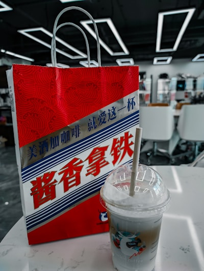 A red and white paper bag with stylized text in Chinese characters, featuring a graphic design with wavy patterns. A plastic cup with a straw, containing a layered iced beverage, is placed on a white table beside it. The background shows an interior setting with modern geometric ceiling lights and white chairs.
