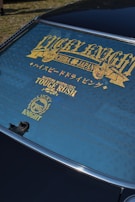 A rear windshield of a car features decorative gold lettering and symbols. The text includes words like 'LUCKY KNIGHT' and 'TOUGE RUSH' with additional Japanese characters. The window is tinted, and the surface has a clean finish.