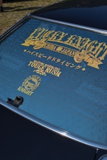 A rear windshield of a car features decorative gold lettering and symbols. The text includes words like 'LUCKY KNIGHT' and 'TOUGE RUSH' with additional Japanese characters. The window is tinted, and the surface has a clean finish.