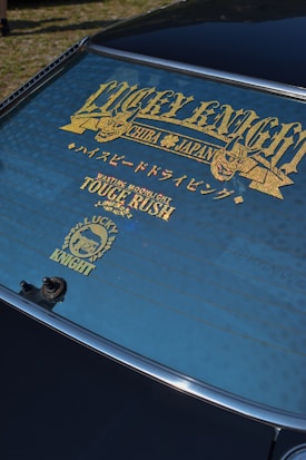 A rear windshield of a car features decorative gold lettering and symbols. The text includes words like 'LUCKY KNIGHT' and 'TOUGE RUSH' with additional Japanese characters. The window is tinted, and the surface has a clean finish.