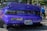 A customized car with a vibrant purple color features various decals and a prominently displayed Japanese license plate. The rear lights are stylish and dark-tinted, and a dual exhaust system is visible. The car is parked on gravel, and in the background, there are people with one person in a wheelchair wearing a blue shirt.
