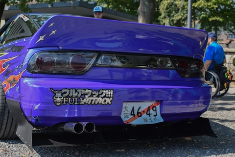 A customized car with a vibrant purple color features various decals and a prominently displayed Japanese license plate. The rear lights are stylish and dark-tinted, and a dual exhaust system is visible. The car is parked on gravel, and in the background, there are people with one person in a wheelchair wearing a blue shirt.