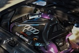 A detailed view of a car engine featuring a large central black carbon-fiber component with silver branding, including 'Audi' and 'V6 Biturbo.' Purple hoses and connections add a distinctive color contrast, while various metallic and plastic components surround the engine.