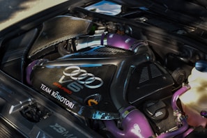 A detailed view of a car engine featuring a large central black carbon-fiber component with silver branding, including 'Audi' and 'V6 Biturbo.' Purple hoses and connections add a distinctive color contrast, while various metallic and plastic components surround the engine.