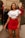A woman stands confidently in a fashion boutique wearing a white crop top with ruffle sleeves and a high-waisted red skirt. Behind her, clothing is displayed on wooden hangers, and a mannequin dressed in a colorful blouse and red shorts is visible. The room is softly lit and has an elegant and stylish atmosphere.
