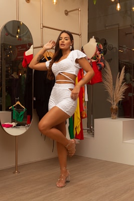 A woman in a stylish white two-piece outfit poses confidently in a boutique setting. The background features a variety of colorful clothing hanging on racks and a mannequin wearing a red and black dress. The environment is warmly lit with decorative light bulbs, and there is a circular mirror and neutral decor elements like a vase with pampas grass.
