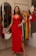 A woman in a red dress stands in a boutique, with clothing on hangers behind her. She is looking confidently at the camera, with her left hand resting on the side of the dress. A full-length mirror reflects part of her image on the left, and a mannequin with colorful clothing can be seen in the background.