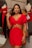 A woman wearing a bright red dress with cut-out details stands in a clothing store. She has long dark hair and is accessorized with earrings. Behind her, there are clothing items hanging on racks and a mirror reflecting part of the scene.
