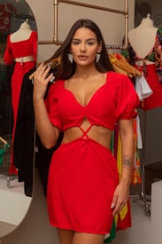 A woman wearing a bright red dress with cut-out details stands in a clothing store. She has long dark hair and is accessorized with earrings. Behind her, there are clothing items hanging on racks and a mirror reflecting part of the scene.