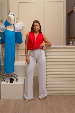 A woman stands confidently beside a mannequin dressed in a blue two-piece outfit. She wears a red sleeveless top with white high-waisted pants and sand-colored heels. The setting appears to be an indoor boutique or fashion store with neutral-colored walls and wood flooring.