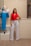 A woman stands confidently beside a mannequin dressed in a blue two-piece outfit. She wears a red sleeveless top with white high-waisted pants and sand-colored heels. The setting appears to be an indoor boutique or fashion store with neutral-colored walls and wood flooring.