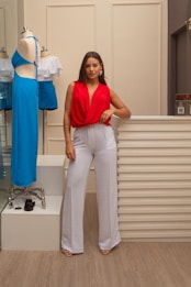 A woman stands confidently beside a mannequin dressed in a blue two-piece outfit. She wears a red sleeveless top with white high-waisted pants and sand-colored heels. The setting appears to be an indoor boutique or fashion store with neutral-colored walls and wood flooring.