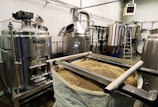 A brewery setup with stainless steel tanks labeled 'Mash' and 'Boil', surrounded by other large tanks and industrial equipment. A bag filled with grains is positioned in the foreground, indicating a brewing or fermentation process.