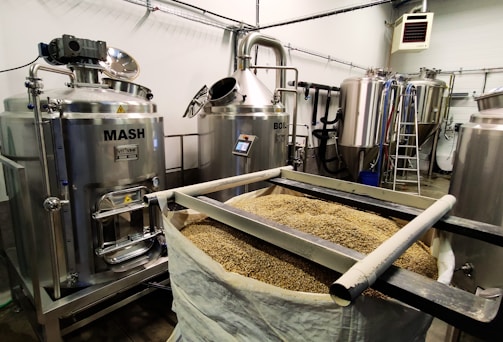 A brewery setup with stainless steel tanks labeled 'Mash' and 'Boil', surrounded by other large tanks and industrial equipment. A bag filled with grains is positioned in the foreground, indicating a brewing or fermentation process.