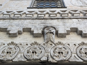 A detailed stone facade features intricate carvings. Geometric and floral patterns adorn the surface, with a central sculpted face that includes a protruding rod from its mouth. The facade has a weathered, textured appearance showcasing its historical character. Above, a window with a decorative grille is partially visible.
