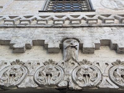 A detailed stone facade features intricate carvings. Geometric and floral patterns adorn the surface, with a central sculpted face that includes a protruding rod from its mouth. The facade has a weathered, textured appearance showcasing its historical character. Above, a window with a decorative grille is partially visible.
