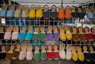 A display of various colorful sandals arranged on a metal rack at an outdoor market. Each row of the rack holds sandals of different colors including blue, yellow, orange, red, green, and more, neatly lined up.