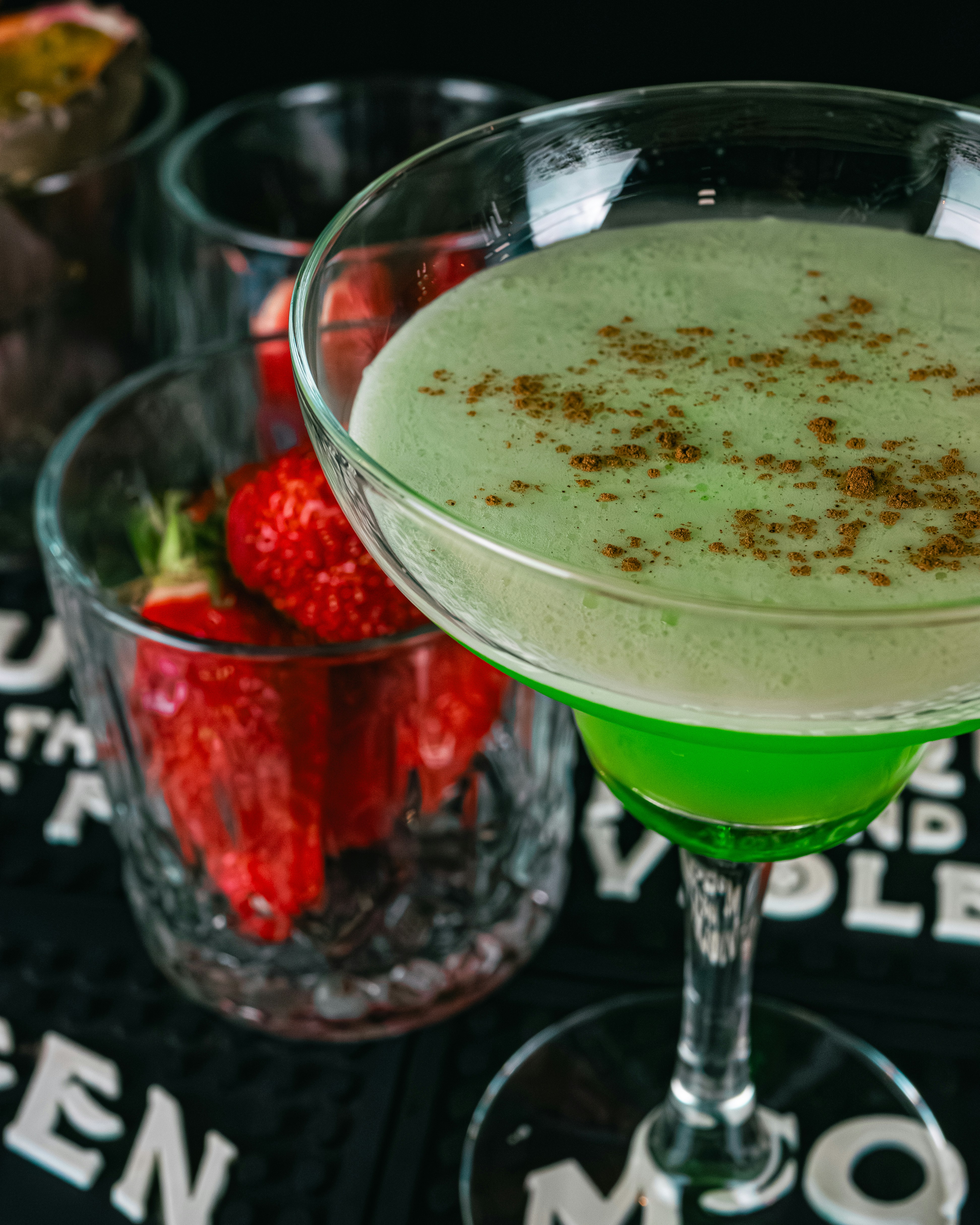 A vibrant green cocktail in a martini glass with a frothy top and a sprinkle of spice. In the background, a glass filled with fresh strawberries adds a contrasting color and texture.