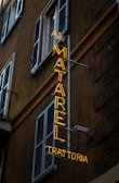 An urban scene featuring a neon sign affixed to the side of a building. The yellow neon sign reads 'AI MATAREL TRATTORIA' and is vertically oriented. The building is dark-colored with multiple shuttered windows, typical of an older architectural style.