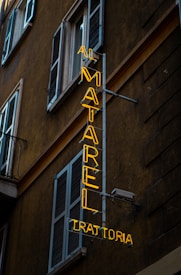 An urban scene featuring a neon sign affixed to the side of a building. The yellow neon sign reads 'AI MATAREL TRATTORIA' and is vertically oriented. The building is dark-colored with multiple shuttered windows, typical of an older architectural style.