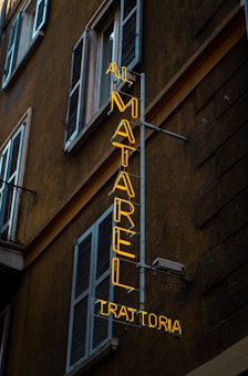 An urban scene featuring a neon sign affixed to the side of a building. The yellow neon sign reads 'AI MATAREL TRATTORIA' and is vertically oriented. The building is dark-colored with multiple shuttered windows, typical of an older architectural style.