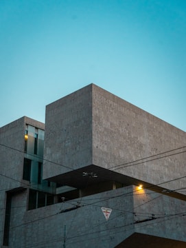 A modern architectural building made of concrete, featuring clean lines and geometric shapes. The structure is set against a clear, blue evening sky. Overhead power lines with a warning sign are visible in the foreground, and a warm light glows from inside one of the building's windows.