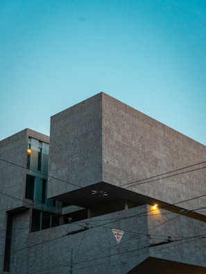 A modern architectural building made of concrete, featuring clean lines and geometric shapes. The structure is set against a clear, blue evening sky. Overhead power lines with a warning sign are visible in the foreground, and a warm light glows from inside one of the building's windows.