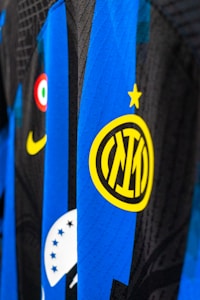 Close-up view of a sports jersey featuring bold vertical stripes in blue and black, adorned with a prominent yellow logo featuring a star on top. Additional elements include a Nike swoosh in yellow, a circular emblem with red, white, and green colors, and a section with black stars on white.