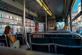 Interior of a city bus with several people seated and looking out the windows. Sunlight illuminates the busy urban street outside, featuring buildings and pedestrians. The scene captures a typical public transport setting.