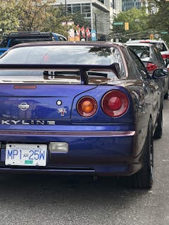 A blue Nissan sports car is parked on a street, with visible features like rear lights and a spoiler. It has a British Columbia license plate. Other cars are visible in the background, along with buildings and trees.