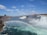 A stunning view of Niagara Falls with mist rising from the powerful waterfalls, creating a vivid rainbow over the river. In the distance, an arched bridge connects the two sides of the river, with buildings visible among trees.