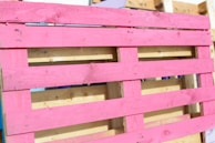 A wooden pallet painted in bright pink, with natural wood color visible in the background. The pallet is leaning against other unpainted pallets, showing a contrast between the vivid pink and the neutral tones of wood.