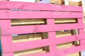 A wooden pallet painted in bright pink, with natural wood color visible in the background. The pallet is leaning against other unpainted pallets, showing a contrast between the vivid pink and the neutral tones of wood.