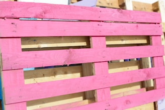 A wooden pallet painted in bright pink, with natural wood color visible in the background. The pallet is leaning against other unpainted pallets, showing a contrast between the vivid pink and the neutral tones of wood.