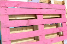 A wooden pallet painted in bright pink, with natural wood color visible in the background. The pallet is leaning against other unpainted pallets, showing a contrast between the vivid pink and the neutral tones of wood.